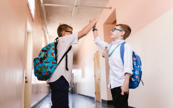 Two boys in school uniform with backpacks, high fiving each other in school corridor 