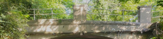 the side of a small rural road bridge over a river, with metal railings alongside the roadway