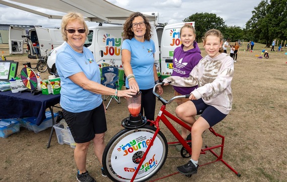 some adults in My Journey t-shirts pose with a child who is making a smoothie with a pedal-powered blender