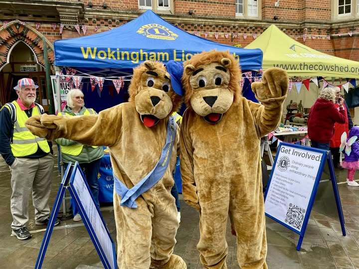 Two people wearing brown lion costumes pose with their arms around each other, with a blue Wokingham Lions Club canopy in the background.