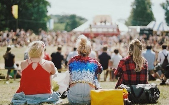Three people sit on the grass with their backs to the camera, facing a stage at an outdoor festival. A large crowd, tents, and flags fill the area.