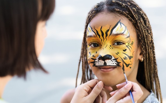 Close-up of a young girl with tiger face paint (orange/yellow, black stripes, white muzzle). She has braided hair and looks at the camera.