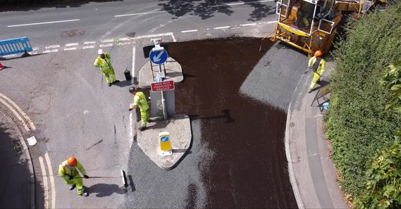 aerial view of a surface dressing vehicle laying stone chippings in a trail of asphalt with men working beside it