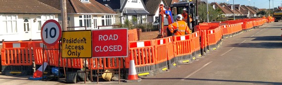 A line of plastic barriers with a "road closed" sign and men using an earth mover behind it