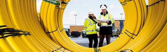 looking through the centre of a big roll of yellow plastic gas pipes, with a man and woman in high vis checking documents