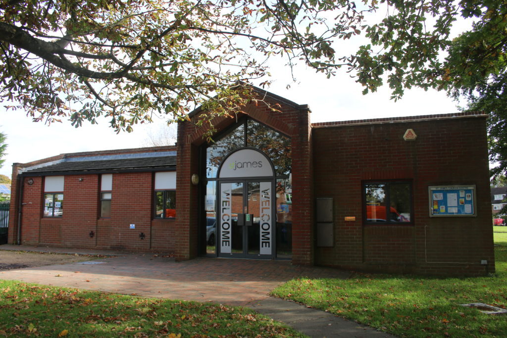 Red-brick St James Church Centre in Woodley, with a glass entrance door marked “Welcome”, set among trees and a paved walkway.