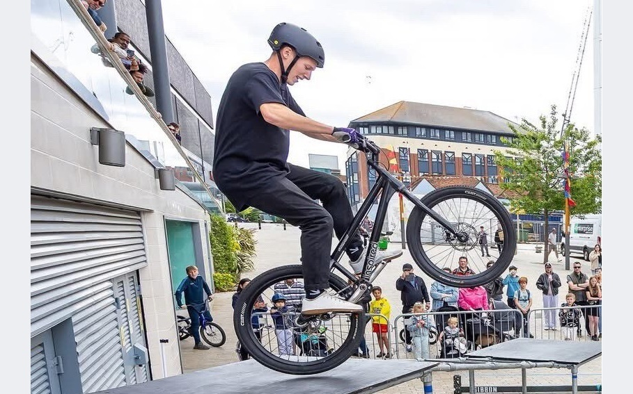 a man does a wheelie on a small BMX bike while poised on the edge of a small ledge, with people watching