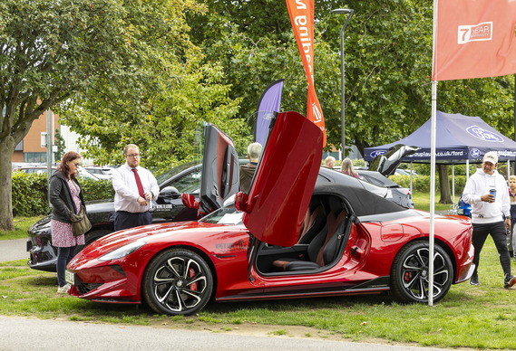 A fancy looking red electric car with its doors lifted up, on a field with other electric cars and people looking at it