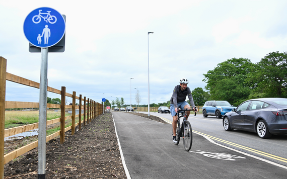 A man on a bicycle rides towards the camera on a shared cycleway alongside a newly opened road