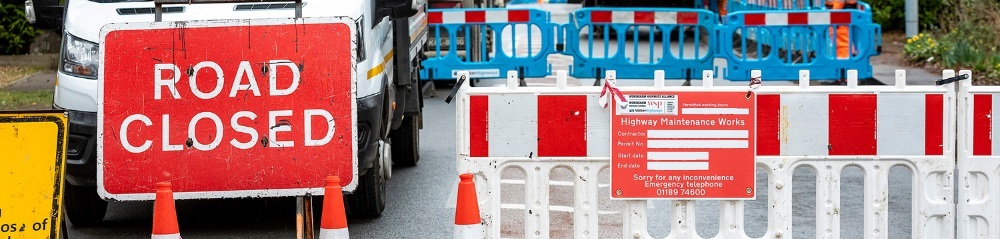 a "road closed" sign in front of a works van, with red barriers closing a road off