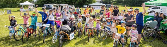 a large mixed group of adults and children sitting on bikes and cheering on the Bikeathon event field
