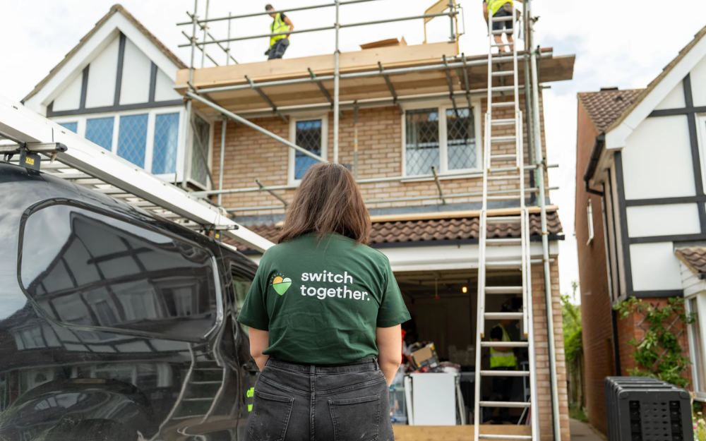 Person in a Switch Together shirt standing outside a house where solar panels are being installed on scaffolding.