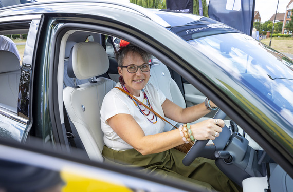 A woman smiles at the camera while sitting in the driving seat of an electric vehicle parked on a field