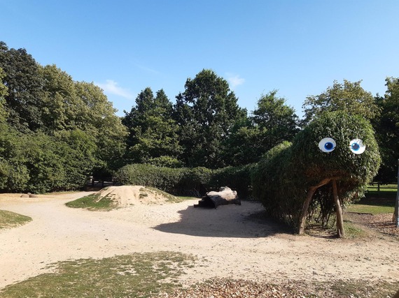 A view of a sculpted living willow that now resembles a caterpillar with googly eyes. Showing the length of the caterpillar and a public park setting