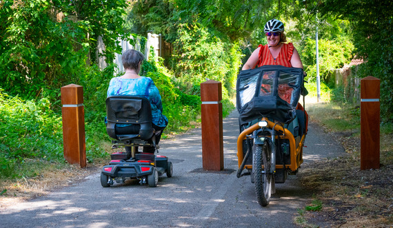 a woman on a mobility scooter and a man on a bike pass in opposite directions on a shared path with a single bollard in the middle