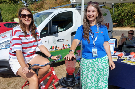 a woman pedals an exercise bike attached to a smoothie maker while a woman from the council holds the lid down
