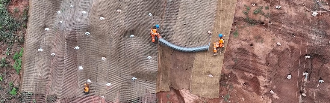 Network Rail workers in orange hi vis abseil down a cliff while driving long steel beams into it