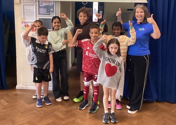a group of children smiling and giving thumbs-up inside a school hall, standing with a My Journey team member