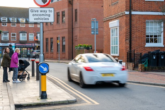 a car, which is blurred due to its speed, drives past two women with a buggy waiting to cross a road in Wokingham town