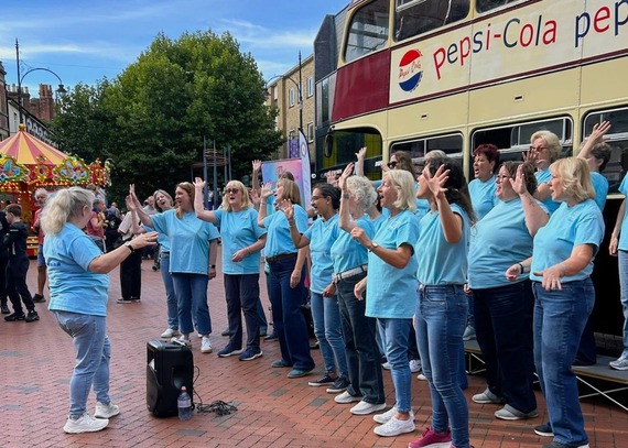 a women's choir in blue T-shirts raise their hands in the air while singing in front of a vintage bus with a Pepsi advert on the side