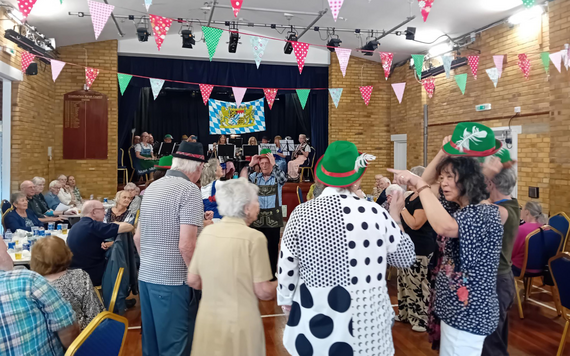 People at an Oktoberfest event, sat at tables around the corners of the room and dancing in the centre