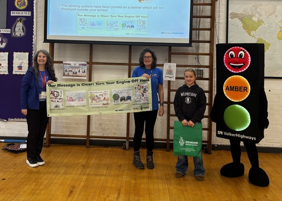 two women and a young schoolgirl pose with a mascot dressed as a traffic signal, holding a banner urging motorists to switch engines off near schools
