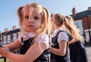 A white, blonde haired Reception aged child in school uniform looking at the camera with her sisters in the background