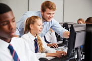 A young male teacher leaning over two secondary age students to point at the screen
