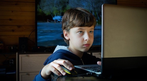 Young boy on a laptop using a mouse. TV in background with a computer game on.