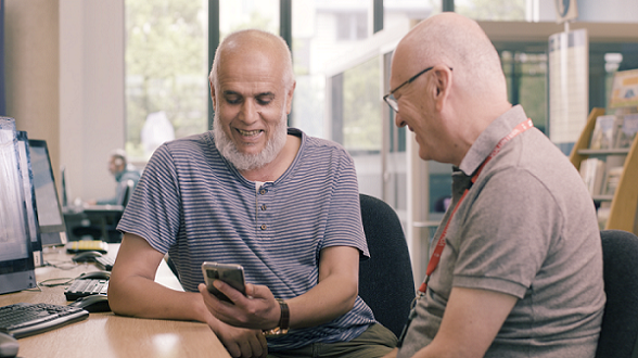 Two males, one wearing a lanyard, sitting in a library looking looking at a smartphone