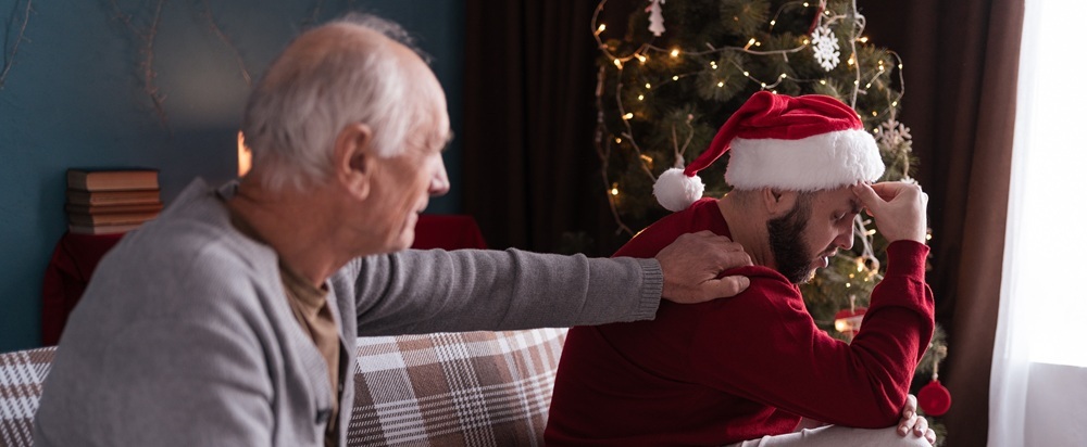 Older man with his hand on a younger male's shoulder who is wearing a Santa hat and red jumper. Both sit on a couch in front of a Christmas tree.