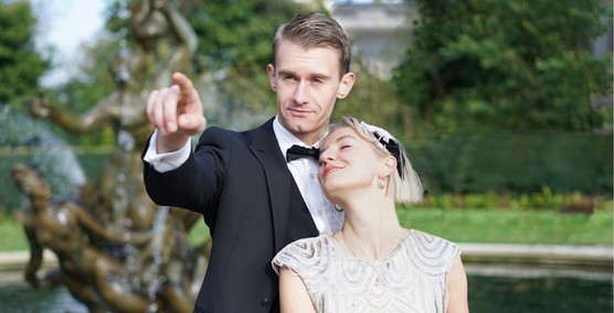 Man in a tuxedo pointing with a lady in a 1920s dress leaning against him