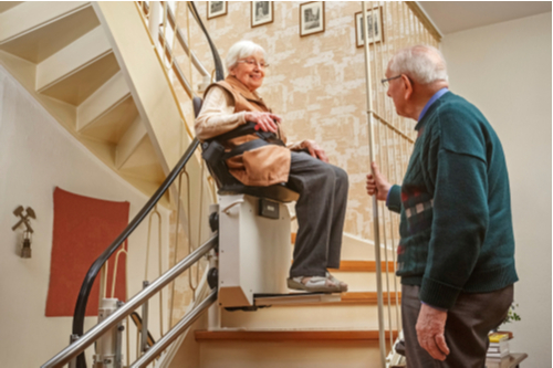 Older lady using a stairlift to go up a curved staircase while a older man stands watching at the bottom of the stairs.