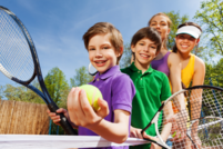 Four children stood behind a tennis net with rackets and balls in their hands
