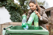 Woman in a brown coat putting three green plastic bottles in a green wheelie bins