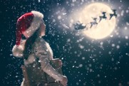 Small girl wearing a santa hat watching father christmas fly in front of the moon on his sleigh.