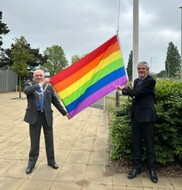 Two gentlemen wearing suits. One has ceremonial chains on. They are holding a rainbow flag.