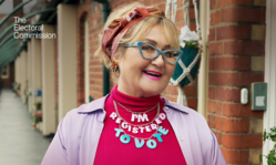 Older lady with headscarf and cat eye glasses, wearing a necklace saying 'I'm registered'