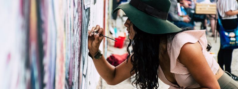 A woman paints a mural onto a wall with a small paintbrush and colourful paints