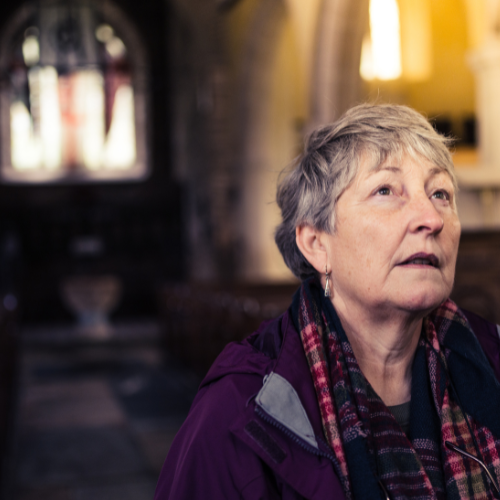 A woman with short hair stands looking into the distance with a beautiful historic building behind and around her