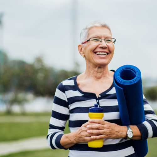 A woman with short grey hair holds a yoga mat and smiles 