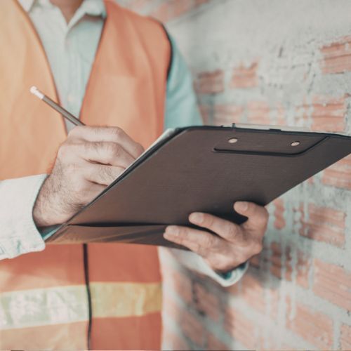 A person in hi-vis writes on a clip board in front of a brick wall