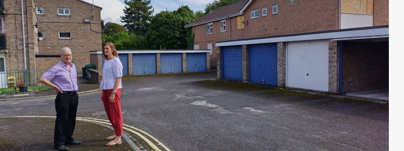 A man and woman stand in a tarmaced area of garages and buildings with boarded windows