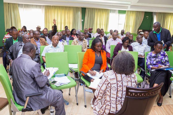 Kenyan cooperative members sit in five rows of green chairs. Two women in the first row take notes. One man raises his hand towards the last rows.