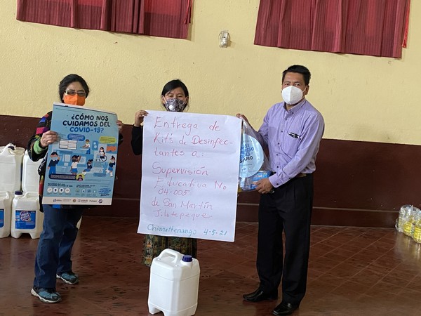 3 school staff in masks hold 2 signs in Spanish, 1 COVID-19 safety flier and 1 to mark when disinfectant kits were delivered to their school.