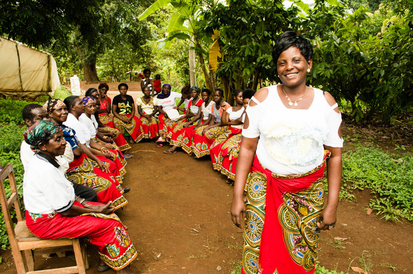 A large group of women in bright red skirts sit on chairs in a semi-circle amid trees. One woman stands in front, smiling at the camera.