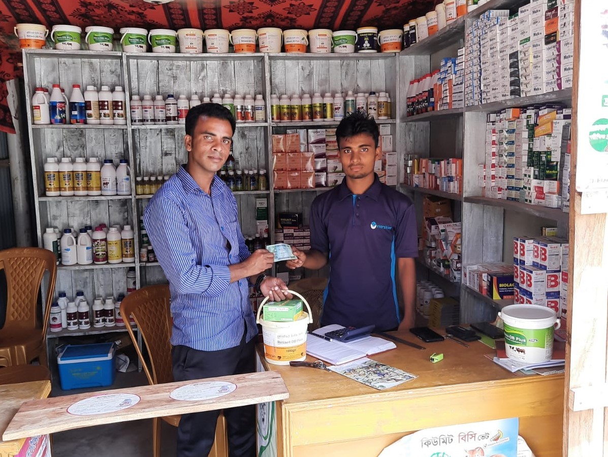 Saiful and Arannayk Foundation staff stand behind Saiful's store’s wood counter. Shelves stocked with animal feed and vet supplies line the walls.