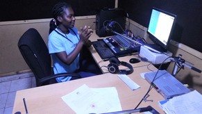 A community librarian claps her hands in the radio station. A microphone and sound equipment to broadcast, and her lesson notes, are on the table.