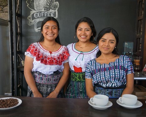 Three young indigenous women, wearing traditional Guatemalan clothing, stand behind the counter of a coffee shop.