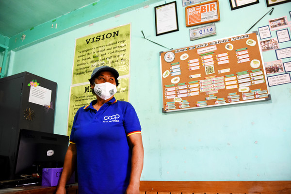 Chairwoman Esperanza in a medical face mask looks to her left, standing in the office of her cooperative with informational posters on the wall.